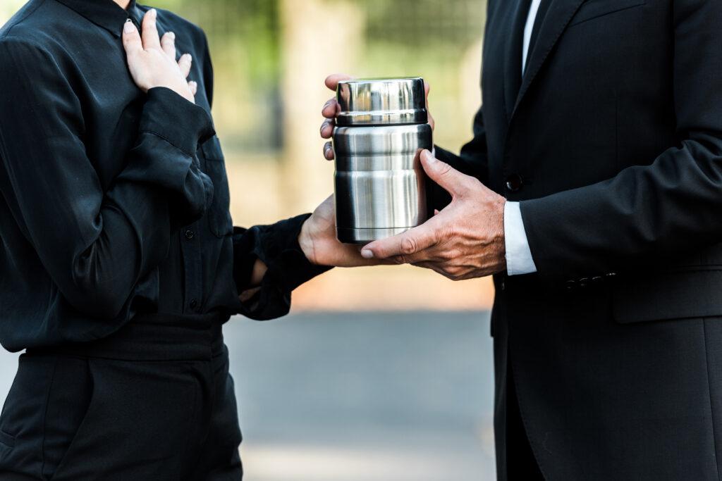 Two people in black suits holding a silver cremation urn between them.