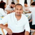 A young man sitting. He is wearing a white shirt, and there are folks in the background.