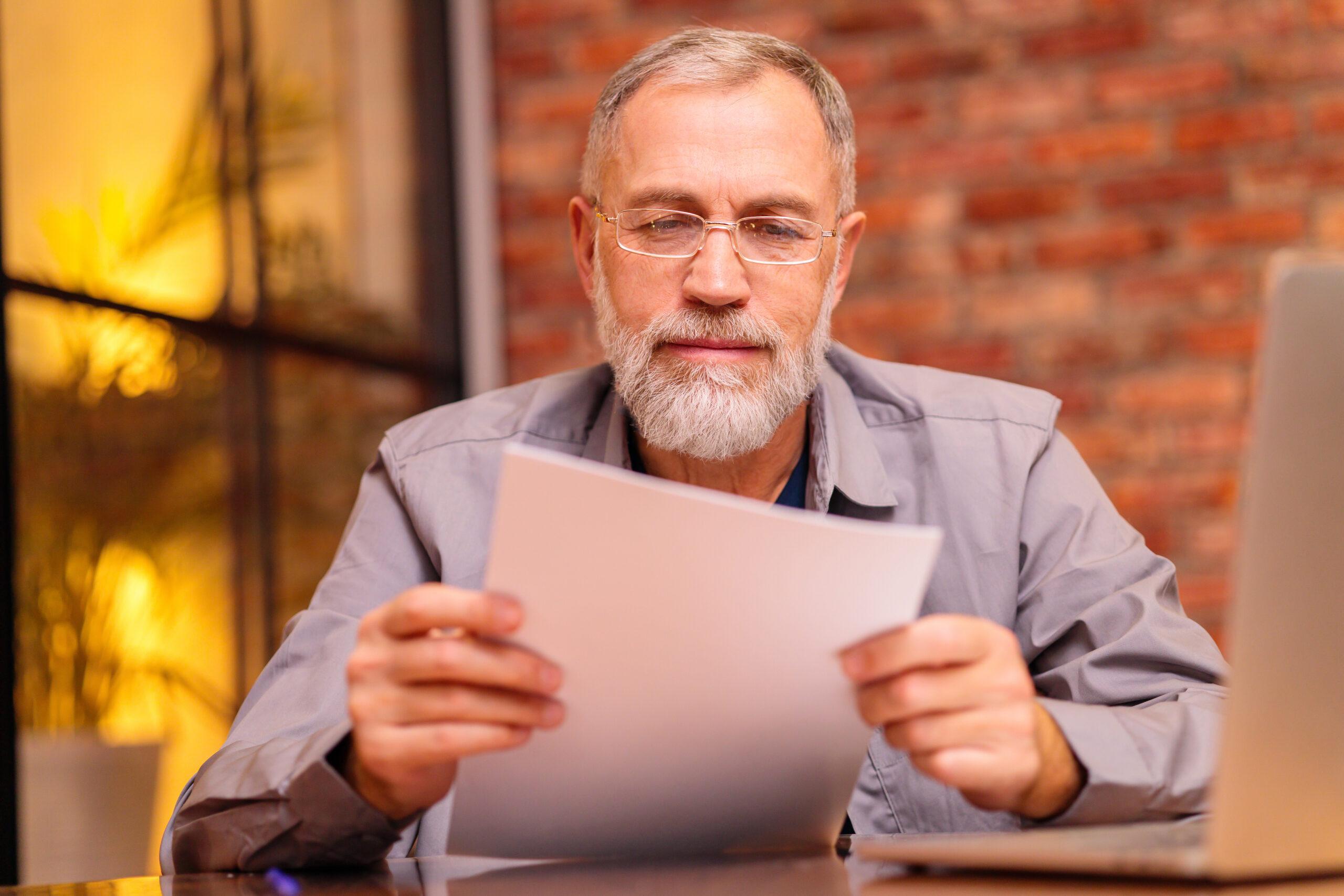 An elderly man reviewing a financial document while seated at a desk.