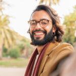 Man with glasses and beard taking a selfie smiling outdoors in the park.