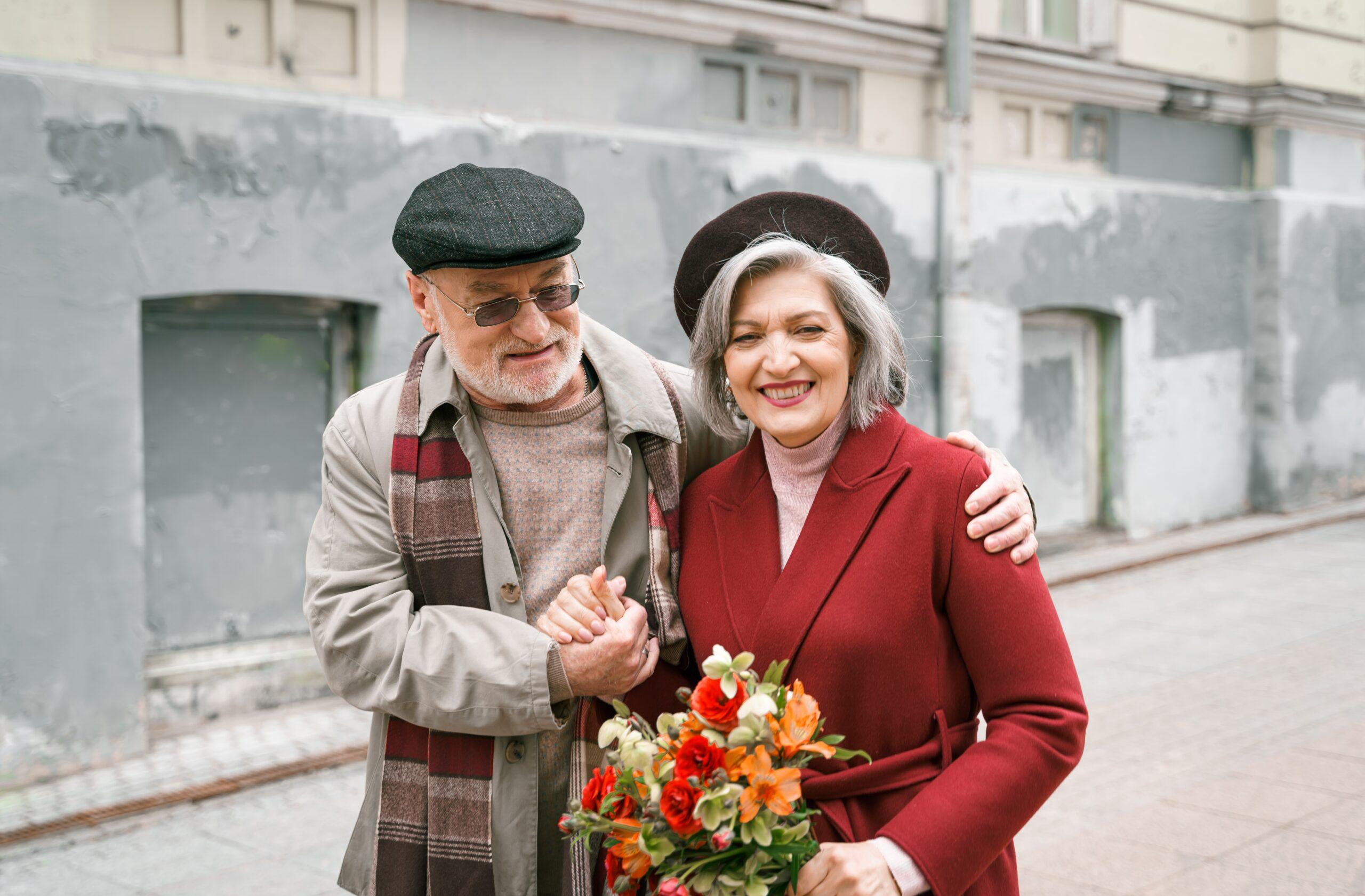 Elderly senior love couple. Old retired man woman together on romantic date.Aged husband wife walking on city street with flowers.Stylish elder hugging people pensioner in red coat.Happy family years