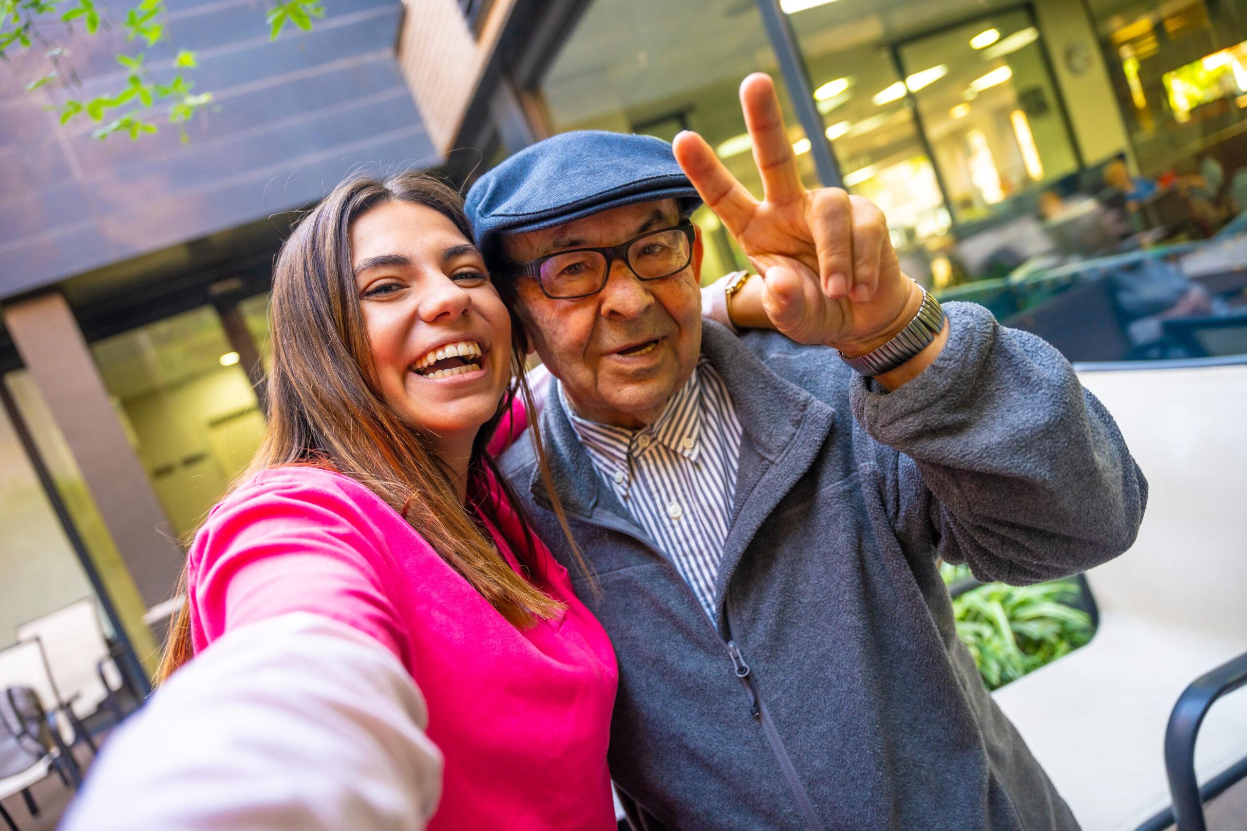 An older man and his granddaughter posing for a selfie photo. He has his hand up in the "peace" symbol.