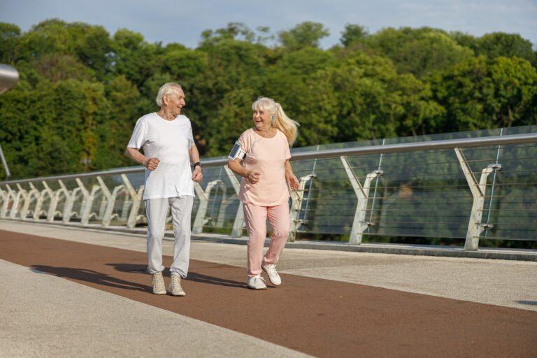 Happy senior man and woman run together along footbridge in summer