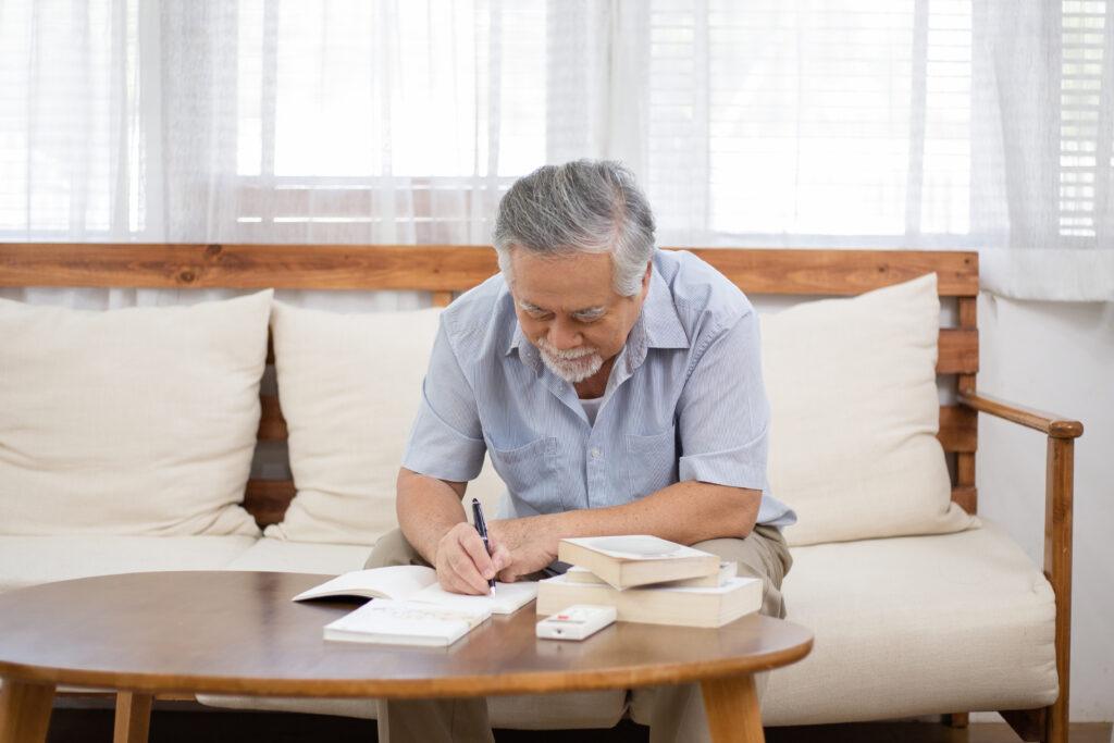 An elderly man wearing a blue shirt and writing in a journal while sitting on a cream colored couch.