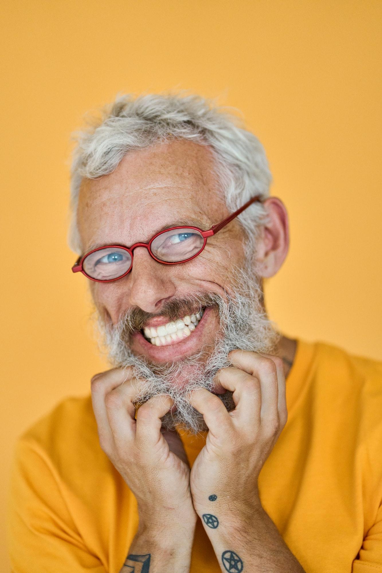Happy funny older hipster man wearing glasses touching beard isolated on yellow.