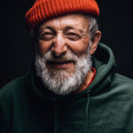 Close up portrait of happy 70-year-old optimist man with smiling wrinkled face, dressed in hipster orange hat and green hoodie, isolated over black background. Positive and cheerful at any age.