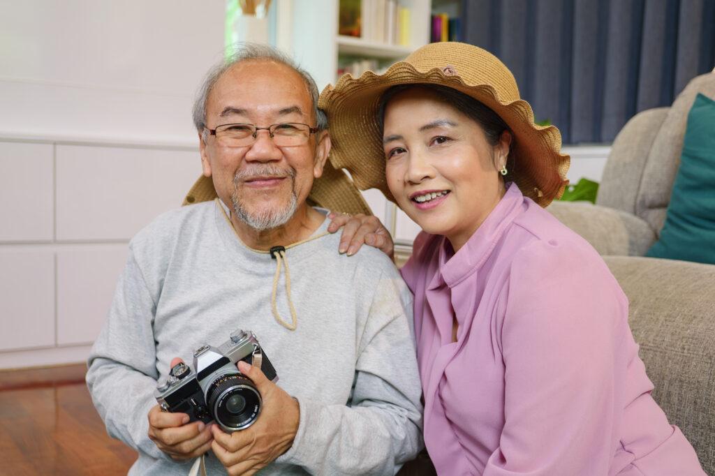 An elderly couple sitting on the floor and smiling at the camera. The woman on the right is wearing a sun hat while the man on the left is holding a vintage camera.