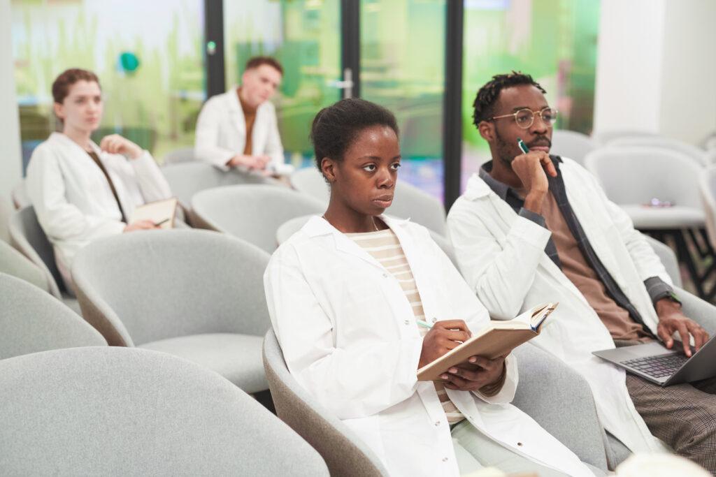 Four doctors taking notes and taking a seminar together. There are two doctors in the foreground and two in the background.