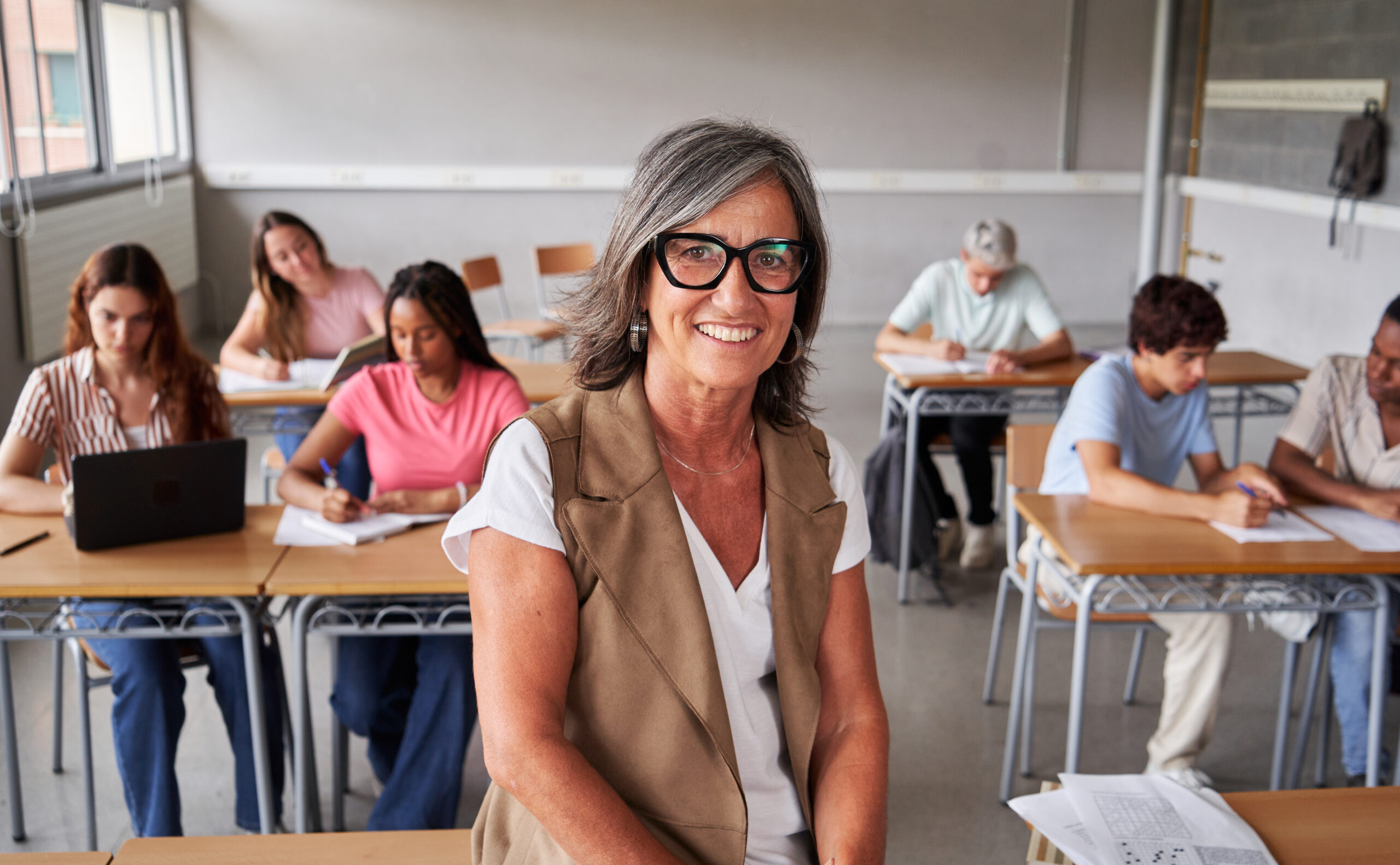 Portrait of a mature teacher at work looks at camera smiling while students study in the background.