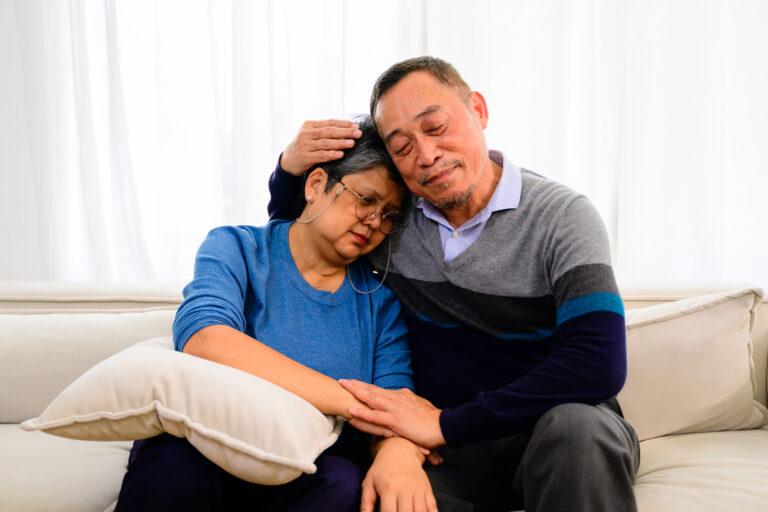 An elderly couple sitting on a white couch together. A man is holding the woman's head and hand while she rests her head against his shoulders.