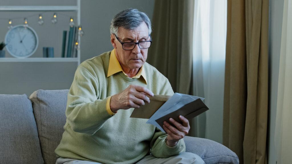 An elderly man with a frustrated look on his face looking at mail.