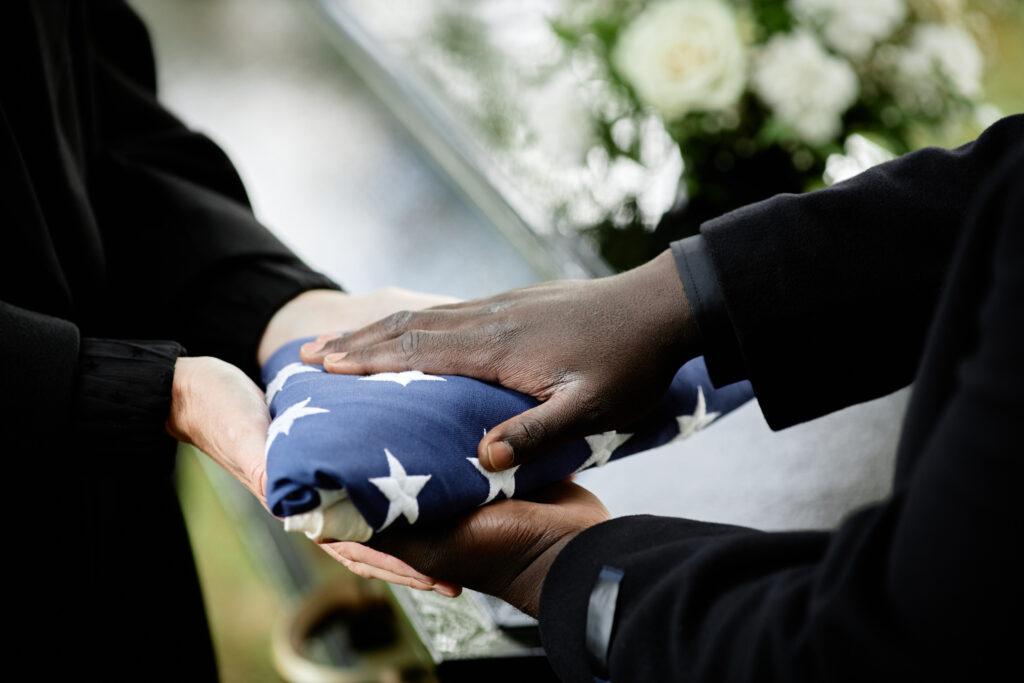 Someone giving another person an American flag with a casket in the background.