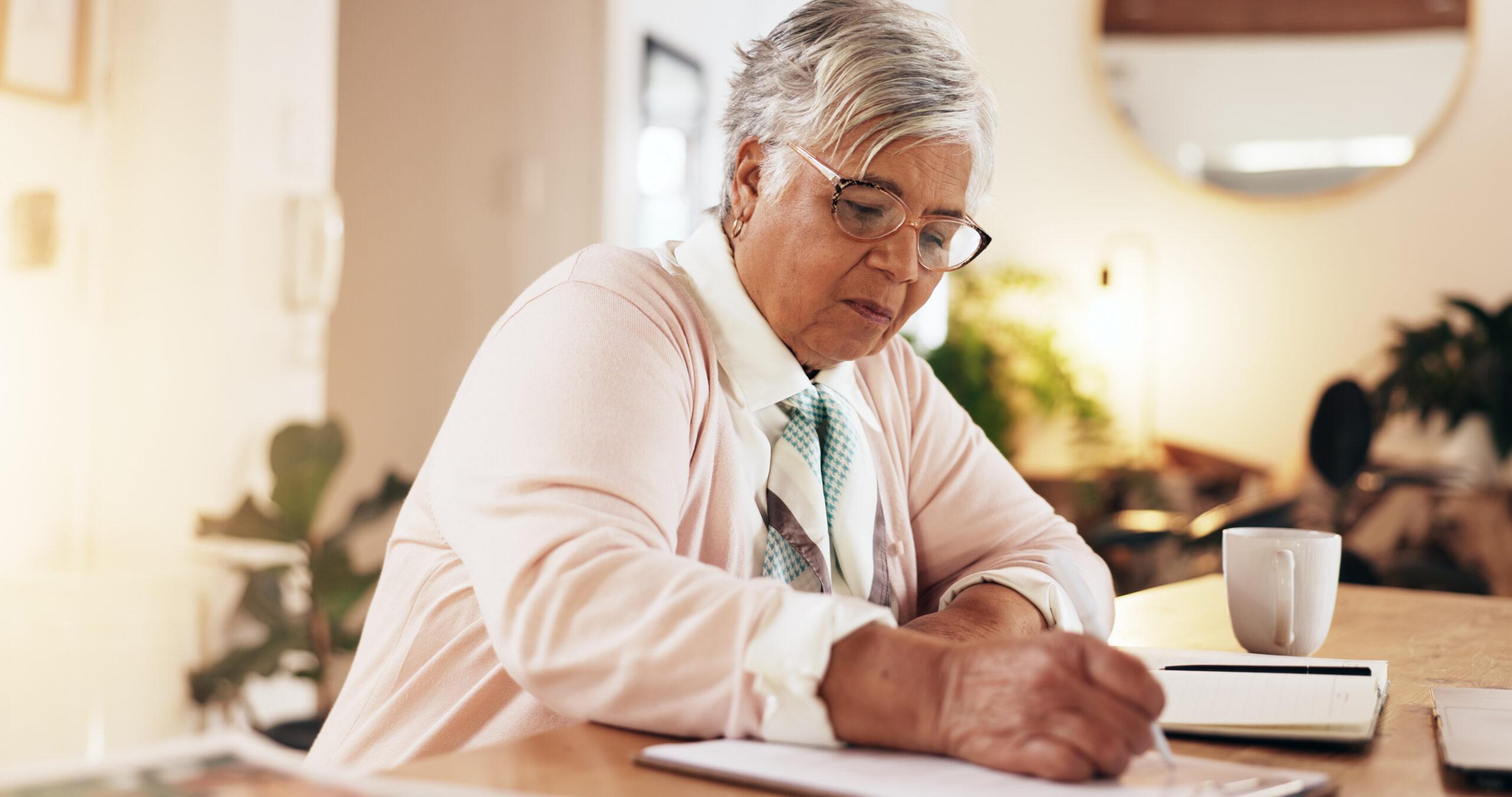 A senior woman wearing glasses writing on paper with a pen