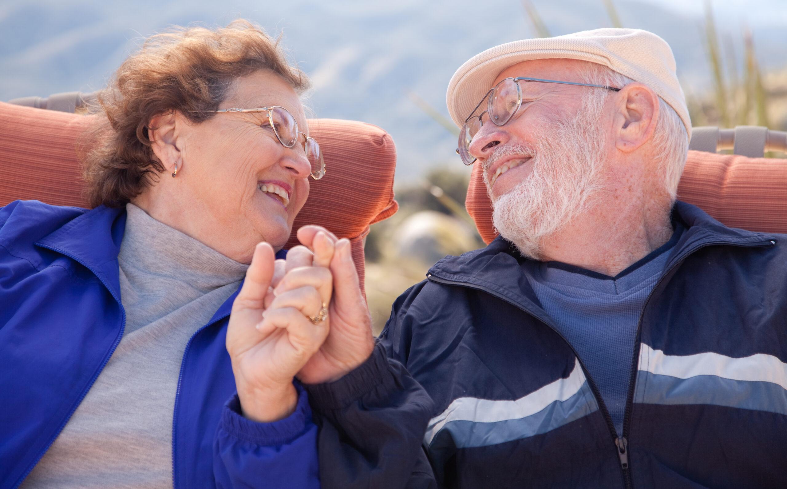 A senior couple seated on recliner chairs holding hands with a landscape behind.