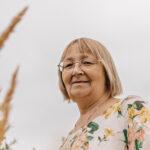 An elderly woman who is standing in a field of tall grass. She is looking into the camera.