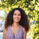 A young woman with curly hair wearing a floral dress outside. The background has tree leaves in the background and a blurry cityscape.