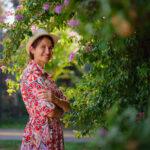 A young woman looking into the camera wearing a red floral dress. She is surrounded by foliage that is blooming purple flowers.