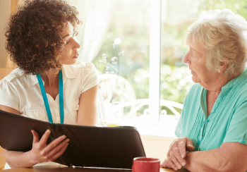 A senior woman is assisted by a caregiver while seated at a kitchen table.