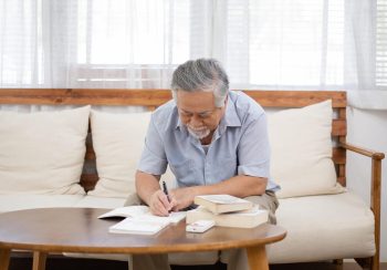 An elderly man wearing a blue shirt and writing in a journal while sitting on a cream colored couch.