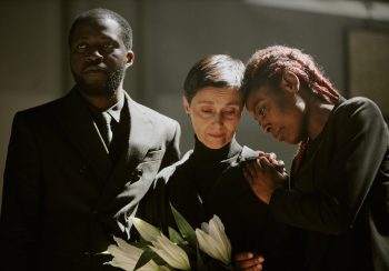 Mournful woman in total black clothes holding madonna lilies surrounded by her African American friends who supporting her and crying