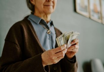 Senior woman holding and counting dollar bills while standing in room wearing a brown sweater with necklace and blue shirt, money is in focus