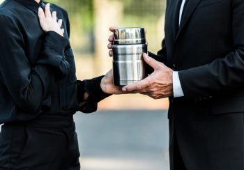 Two people in black suits holding a silver cremation urn between them.