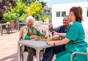 An elderly man and woman, along with a nurse, happily engage in a children's game for hand exercises on a sunny day at a geriatric patio.