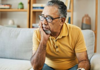 Face, depression and alzheimer with a sad old man on a sofa in the living room of his retirement ho.