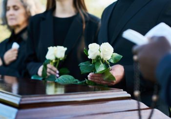 Focus on two fresh white roses held by mourning man in black suit during funeral service while standing by coffin against his daughter and wife