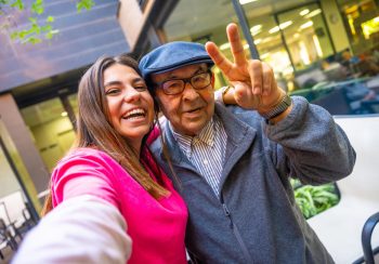 An older man and his granddaughter posing for a selfie photo. He has his hand up in the "peace" symbol.