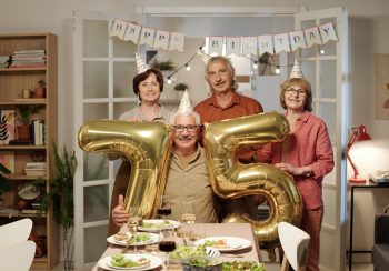 Group of senior friends holding inflatable numbers showing age of one of them while standing in front of festiv table served with homemade food