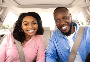 Front portrait of smiling beautiful African American couple sitting in modern luxury car, happy excited young man and woman posing looking at camera, enjoying road trip or buying new car