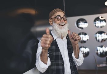 Happy aged male in glasses and hat with gray beard showing thumbs up gesture while looking at camera and smiling during break in office