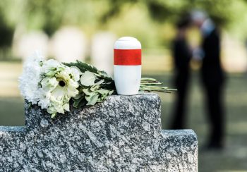 A cremation urn on top of a gravestone beside some flowers.