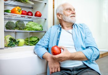 Portrait of a thoughtful senior man sitting with apple near the refrigerator full of healthy food at home