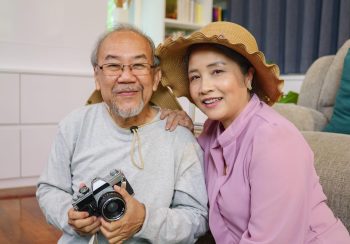 An elderly couple sitting on the floor and smiling at the camera. The woman on the right is wearing a sun hat while the man on the left is holding a vintage camera.