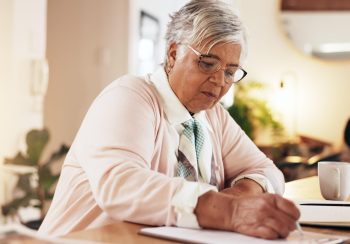 A senior woman wearing glasses writing on paper with a pen