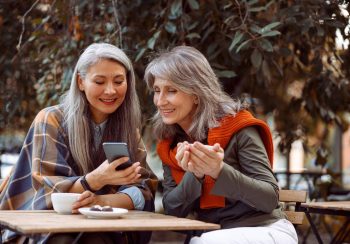 Asian lady shows photos on mobile phone to silver haired friend with cup of tea sitting at small table in street cafe