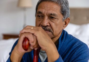 Thoughtful senior biracial man sitting and holding walking stick at home. Medical services, check up, home visit, healthcare, wellbeing and senior lifestyle, unaltered.
