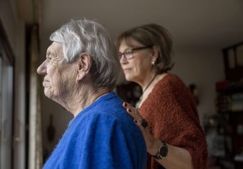 Two elderly friends standing looking out of the window consoling one another